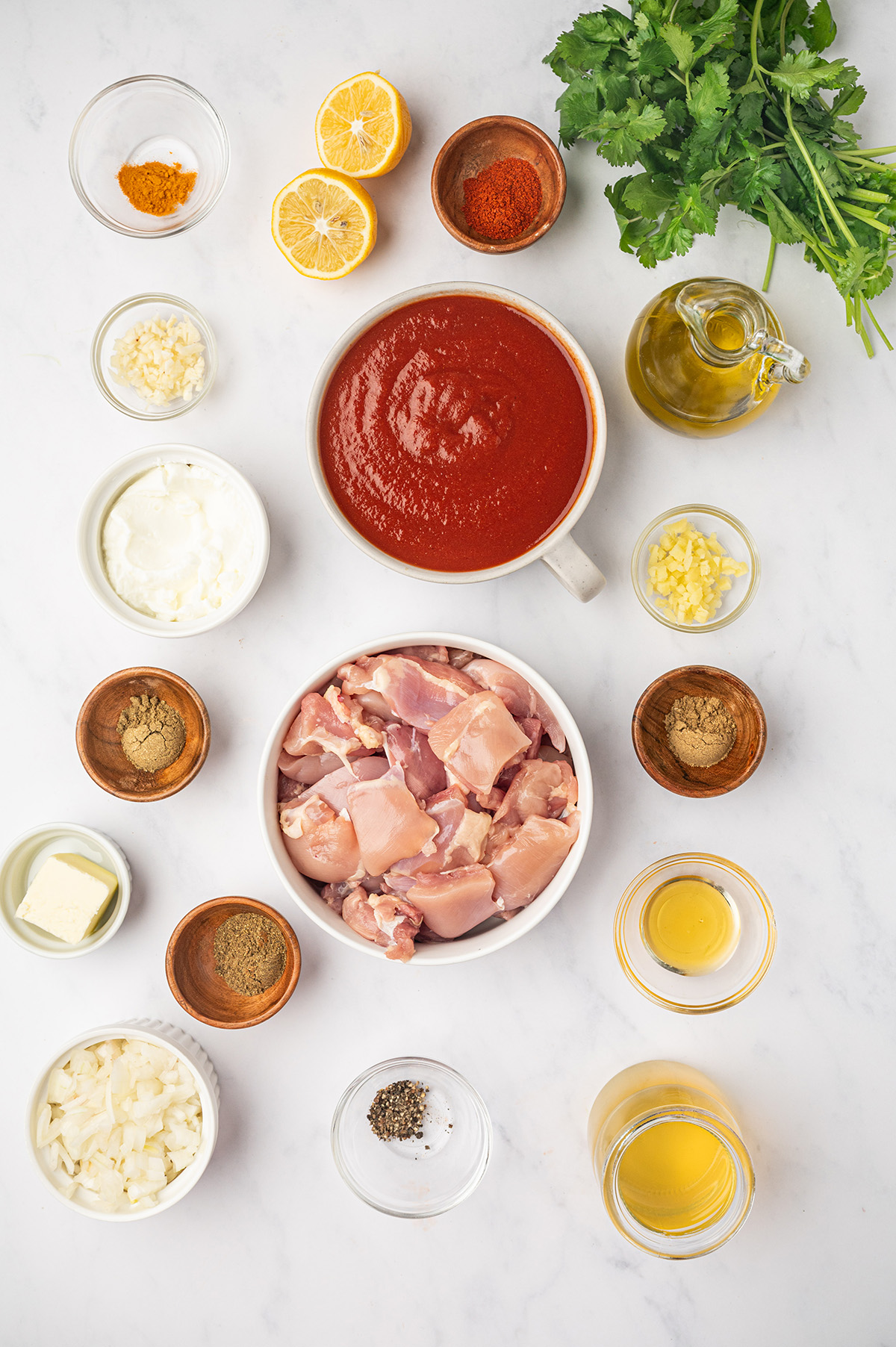 A flat-lay of ingredients for low sodium butter chicken on a white surface, including chicken, tomato sauce, warm spices, and unsalted butter.