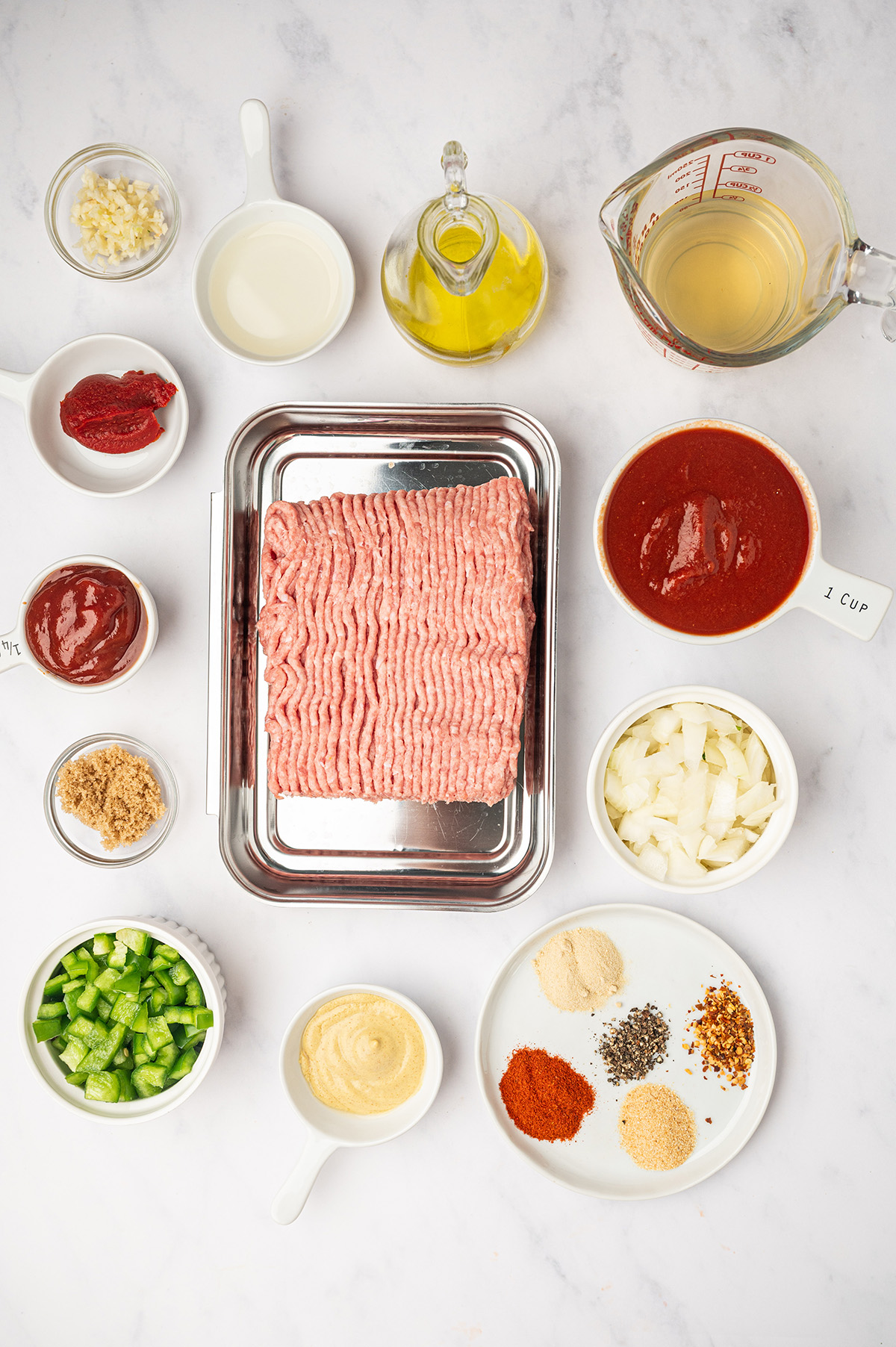 A flat-lay of ingredients for low sodium sloppy joes on a white surface, featuring ground meat, tomato sauce, unsalted butter, chicken broth, and a homemade seasoning mix.