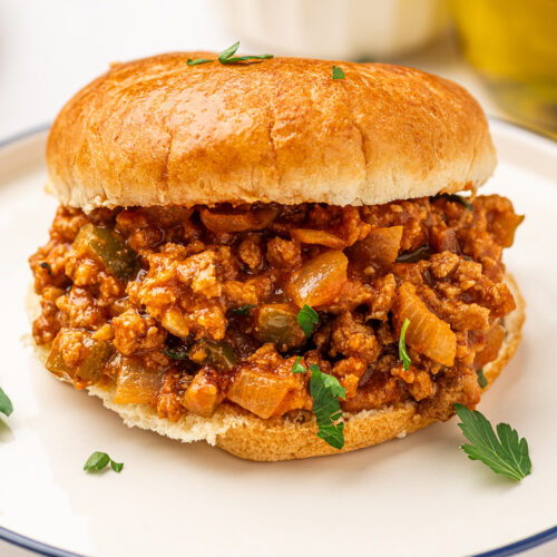 A low sodium sloppy joes sandwich on a toasted bun, garnished with fresh parsley and served on a white plate.