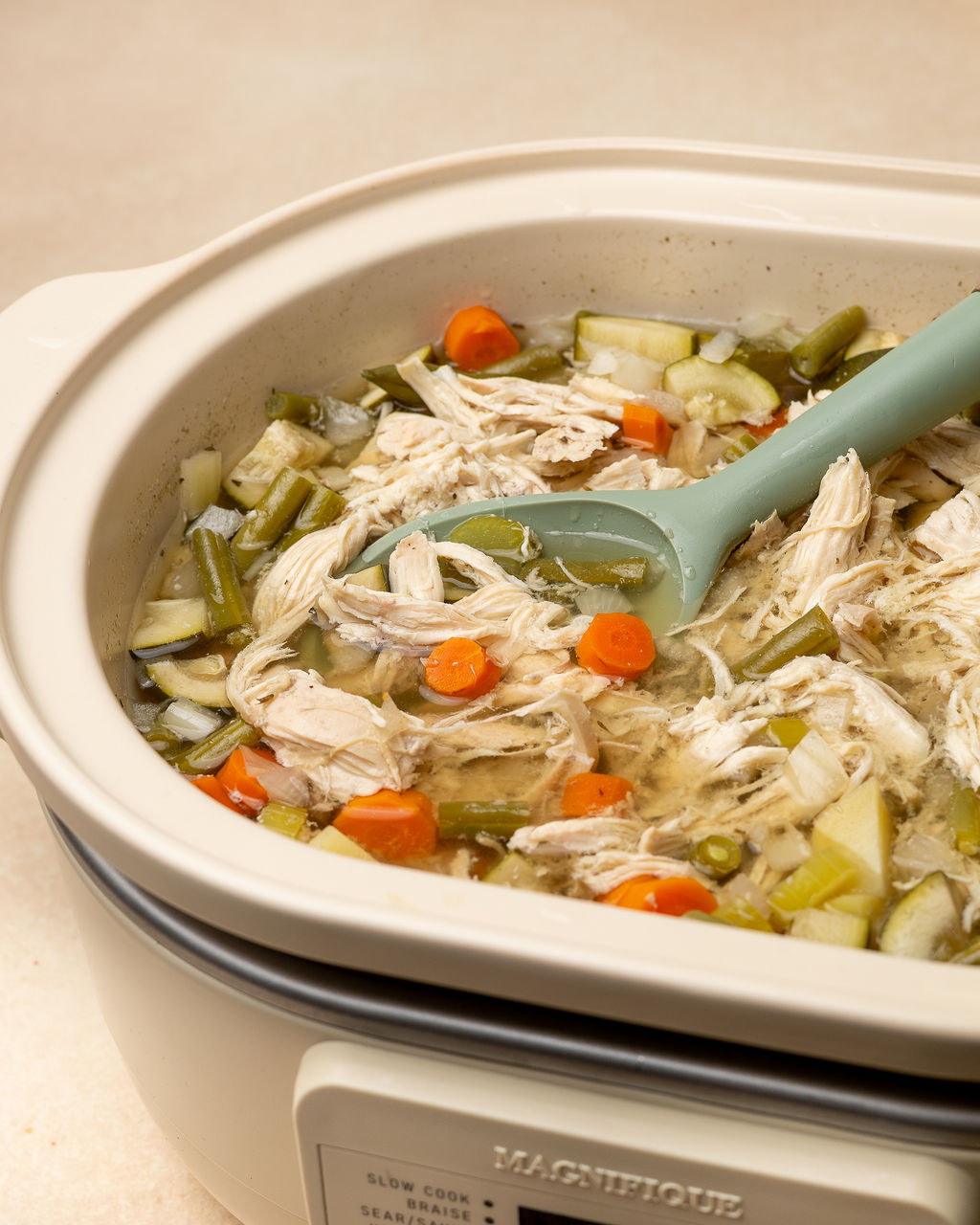 A serving spoon dipping into a slow cooker of chicken and vegetable soup.