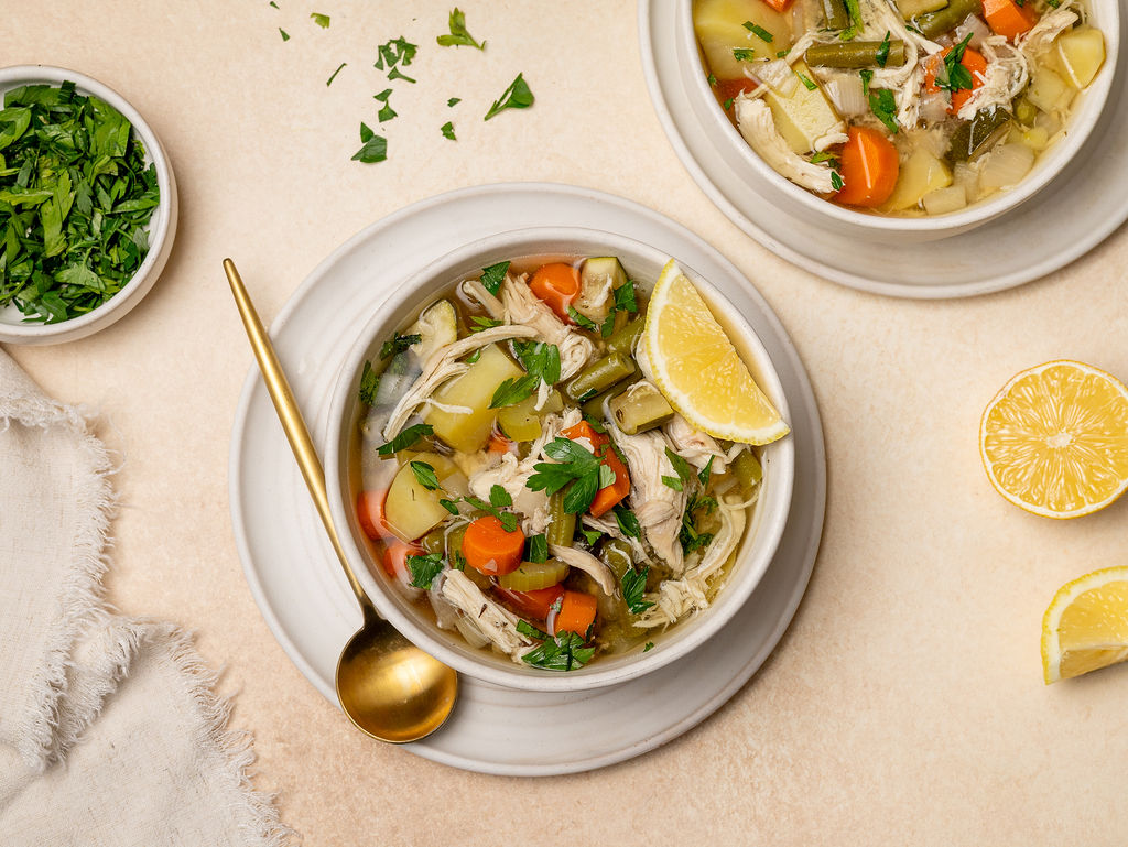 Two servings of chicken and veggie soup with chopped cilantro and lemon wedges.